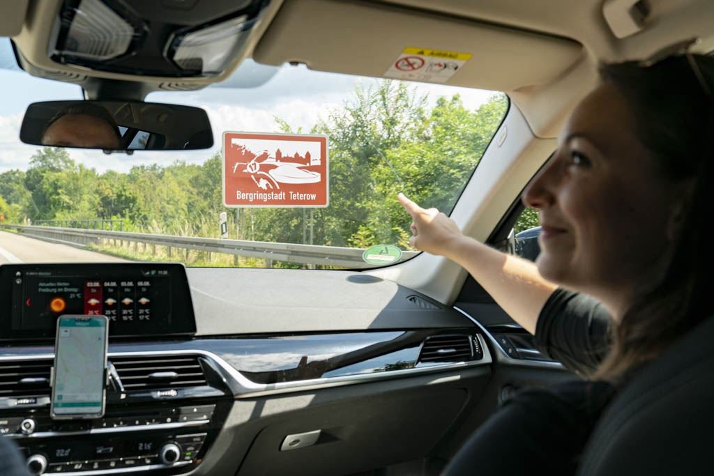 Ein Mann und eine Frau fahren auf der Autobahn. Sie zeigt auf ein braunes Schild mit der Aufschrift 